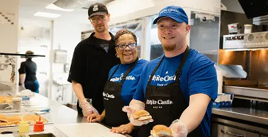 Three White Castle employees smile behind the counter, with one holding two sliders in a restaurant kitchen.