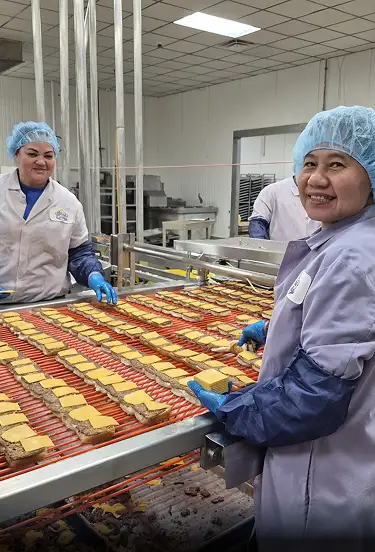 White Castle team members in white & blue uniform place cheese on sliders along a conveyor line in a food processing facility.