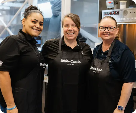Three White Castle employees smile together in the restaurant kitchen wearing black aprons.