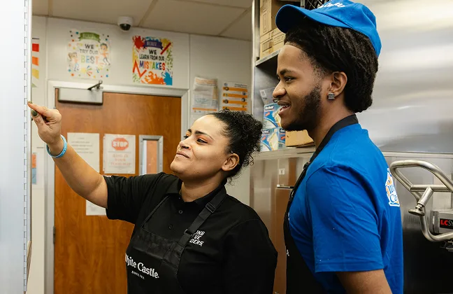 Two White Castle employees smile while reviewing information posted on a kitchen wall.