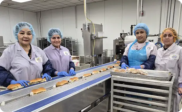 Four food production workers stand beside a conveyor of slider buns in a White Castle bakery facility.