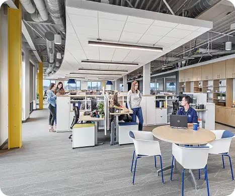 Employees collaborate in a modern open office with workstations, meeting space, and shared seating.