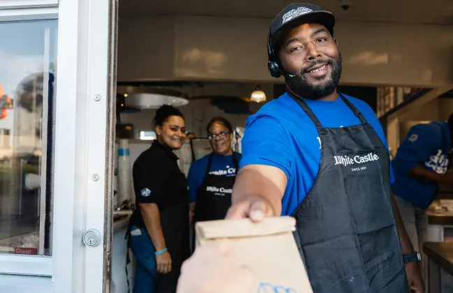 White Castle employee hands a takeout order through the drive-thru window as coworkers look on.
